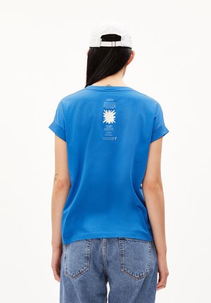 Persona con cabello largo y oscuro que lleva una gorra blanca, camiseta azul con texto gráfico blanco en la espalda y vaqueros azules, de espaldas.