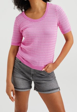 Kobieta ubrana w różowy krótki sweter z dzianiny i szare szorty jeansowe, stojąca z jedną ręką w kieszeni na jednolitym tle.