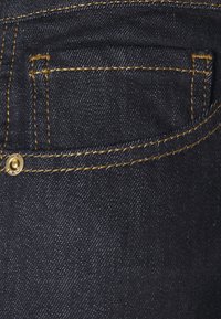 Poche en denim bleu foncé avec surpiqûres jaunes et un petit rivet métallique dans le coin, montrant le tissu texturé et les détails des coutures.