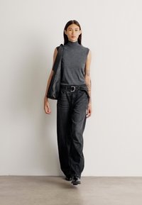 Siva brezrokavnik s high neck, v kombinaciji s prostornimi temnimi jeans hlačami. Model nosi črno torbo, kar prikazuje preprosto, moderno obleko.