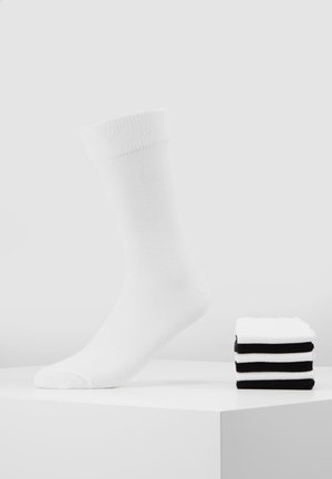 White cotton sock with a ribbed cuff, next to a stack of five black and white socks. Smooth texture and medium height design.