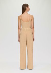 Beige Plissees-Jumpsuit mit dünnen Trägern, einem runden Rückenausschnitt und weitem Bein-Design. Strukturierter Stoff mit vertikalen Linien, taillierte Passform.