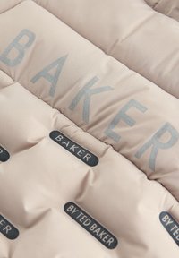 Puffatakki pehmeästä persikanvärisestä kankaasta, jossa on ommeltuja osia. Sisältää hopeiset "BAKER"-kirjaimet ja mustat "BY TED BAKER"-tunnisteet.