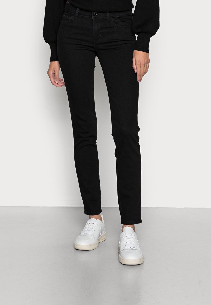 Personne portant un jean noir slim et des baskets blanches, debout devant un fond clair uni.