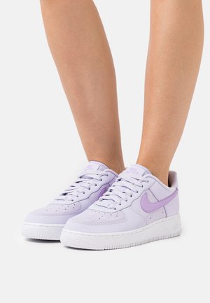 Personne portant des baskets Nike Air Force 1 lavande avec des semelles blanches, debout sur un fond gris clair uni.