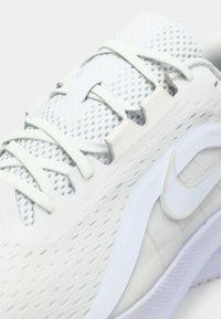 Λευκό αθλητικό sneaker με επάνω μέρος από διχτυωτό υλικό, επίπεδες ανοιχτό γκρί κορδόνες, υφή στην σόλα και μια διακριτική λογότυπη λεπτομέρεια στο πλάι.