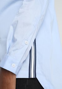 Camicia azzurra a manica lunga con polsino bottone, caratterizzata da un dettaglio a strisce verticali contrastanti in blu navy e bianco lungo la cucitura laterale.