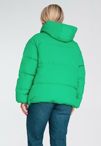 Zielona kurtka puchowa z kapturem, o pikowanej konstrukcji i prostokątnym kroju, zestawiona z niebieskimi dżinsami. Widoczne detale przeszycia.