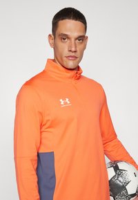 Haut de sport orange à demi-zip avec un panneau latéral bleu marine, présentant une texture lisse et le logo Under Armour en blanc. Un ballon de soccer est tenu dans une main.