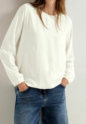 Mujer con una blusa blanca de manga larga y suelta con puños elásticos y jeans vaqueros azules con las manos en los bolsillos.