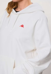 Mujer con sudadera blanca con capucha y un logo rojo de dos siluetas sentadas en el pecho, una masculina y una femenina, espalda con espalda.