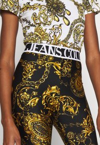 Weißes T-Shirt mit komplexem goldenen Muster, kombiniert mit schwarzen Leggings, die ein ähnliches vergoldetes Design aufweisen. Elastischer Bund mit dem Text "JEANS COULTURE".