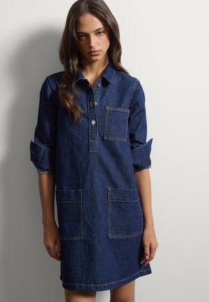 Giovane donna con capelli lunghi e scuri indossa un vestito di jeans blu scuro con tre tasche frontali e maniche arrotolate su uno sfondo semplice.