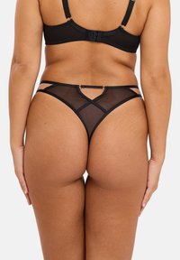 Ensemble de lingerie en mesh noir comprenant un bas thong avec un design de découpes complexe et des bretelles fines, rehaussé par des accessoires en métal doré.