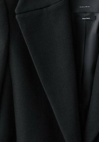 Zwarte blazer met gestructureerde stof, scherpe revers en een gladde voering. Label luidt "Italiaanse stof." Klassiek getailleerd ontwerp.