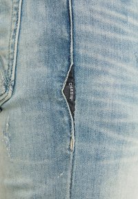 Jean en denim bleu clair avec une petite étiquette en tissu noir indiquant "CHASIN'" cousue dans une couture latérale.