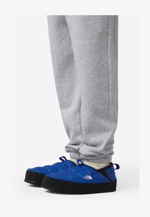 Chaussures à enfiler bleues isolées avec semelles en caoutchouc noires et marquage. Portées avec un pantalon de survêtement gris, présentant un design rembourré et une hauteur à la cheville.