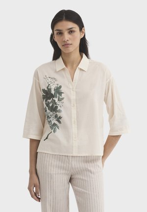 Frau trägt ein cremefarbenes Hemd mit Knöpfen und grünem Blumenmuster auf einer Seite sowie hellbeige Hose mit Nadelstreifen, steht vor einem schlichten Hintergrund.