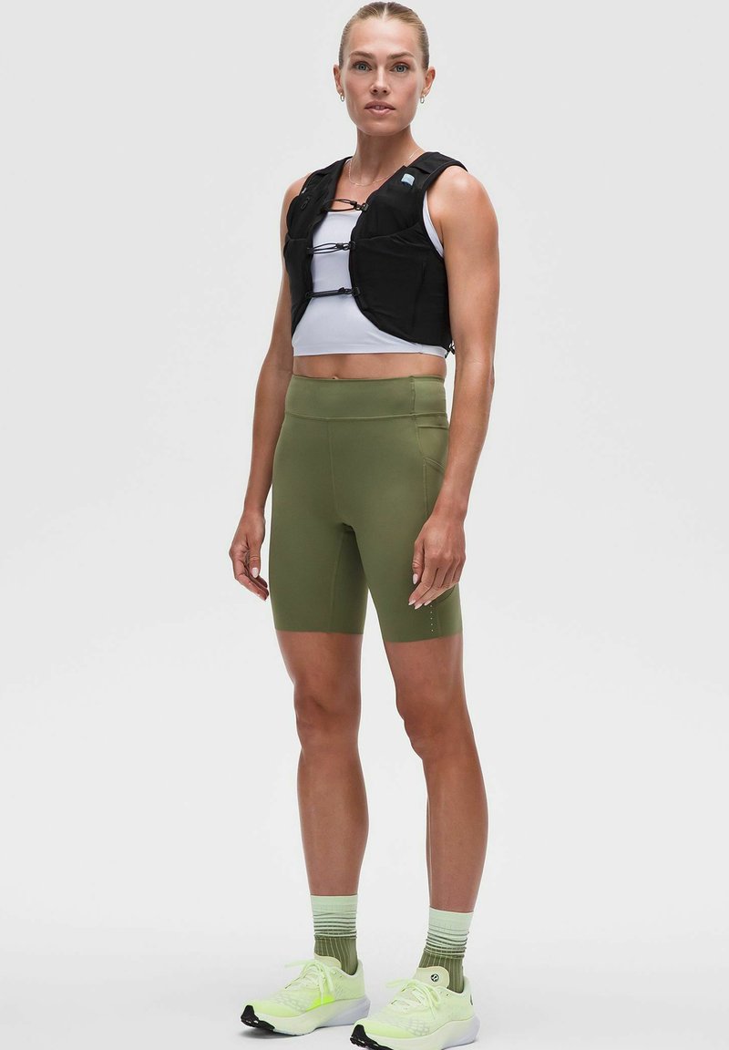 Gilet pesante nero con cinghie regolabili, abbinato a un top corto bianco e pantaloni bike verde. Presenta sneaker verde chiaro e calzini a righe.