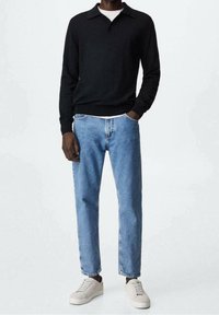 Homme portant un polo noir à manches longues, un jean droit bleu clair, des baskets blanches, se tenant avec une main dans la poche contre un fond uni.