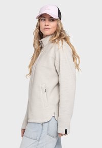 Hellgraue Fleecejacke mit hohem Kragen, seitlichen Taschen und abgerundetem Saum; getragen mit einer pinken und schwarzen Mütze.