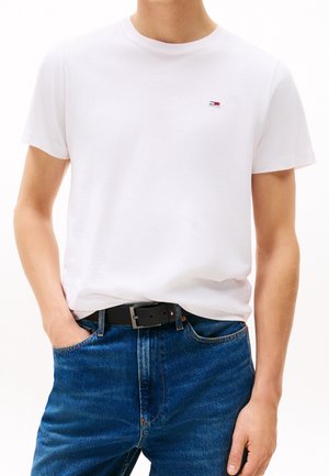 Mężczyzna w prostym białym t-shircie z małym logo na piersi, wpuszczonym w niebieskie dżinsy z czarnym paskiem, z jedną ręką w kieszeni.
