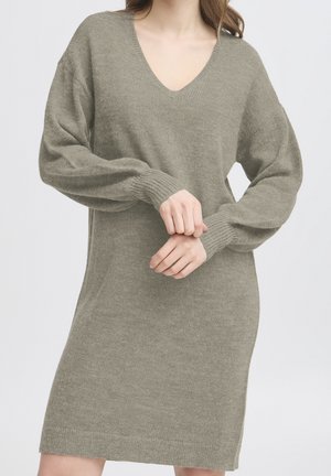 Femme portant une robe pull marron clair en maille à manches longues et col en V, les mains jointes à la taille devant un fond clair uni.
