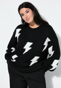 Frau trägt einen schwarzen Pullover mit weißen Blitzmustern und steht mit den Händen in den Taschen vor einem weißen Hintergrund.