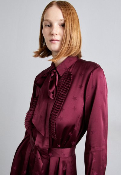 El vestido de seda burdeos cuenta con un lazo, detalles plisados en el torso y patrones de estrellas. El diseño incluye mangas largas y una cintura ajustada.