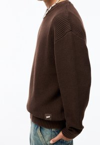 Ruskea neulottu villapaita, jossa on ribbineulos, pyöreä pääntie ja levenevät olkapäät. Alareunassa pieni logoplätsi.