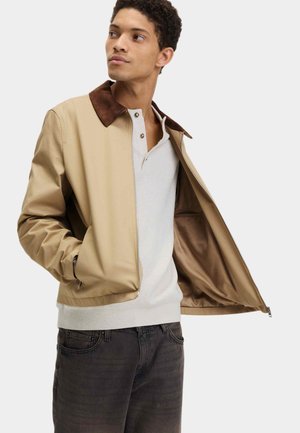 Junger Mann trägt eine beige Jacke mit braunem Kragen über einem weißen Henley-Pullover und dunklen Jeans, hält die Jacke geöffnet.