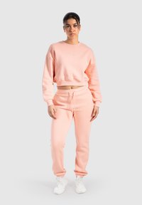 Lys pink cropped sweatshirt med ribbede manchetter og matchende sweats, der har elastisk talje og afsluttede ankler, båret med hvide sneakers.