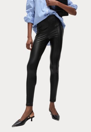 Person trägt schwarze Leggings aus Kunstleder, schwarze spitze Slingback-Pumps und ein blau gestreiftes Hemd und hält eine schwarze Handtasche.