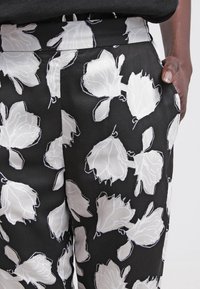 Schwarze Stoffhosen mit einem weißen Blumenmuster und konturierten Details. Der Stoff hat eine glatte Textur und besitzt auf beiden Seiten Taschen.