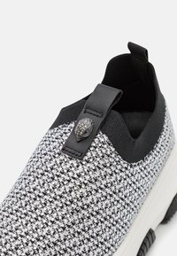 Schwarz-weißer Slipper mit einem strukturierten Strickobermaterial, das eine dekorative schwarze lederne Lasche und einen mit Silber-Nieten verzierten Akzent aufweist. Gummisohle.