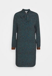 Långärmad klänning i mörkt teal tyg med blommönster, som har en fluga i kragen och leopardtryck på ärmsluten.