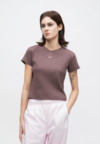 Νεαρή γυναίκα με καστανά μαλλιά, φορώντας ένα μωβ κοντό μπλουζάκι της Nike και ανοιχτό ροζ παντελόνι της Nike, στέκεται με το ένα χέρι στην τσέπη, σε ουδέτερο φόντο.