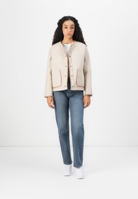 Beige, quiltad jacka med rund halsringning och tryckknappar, med två framfickor med kontrasterande beige kant. Bärs över blå jeans och vita sneakers.
