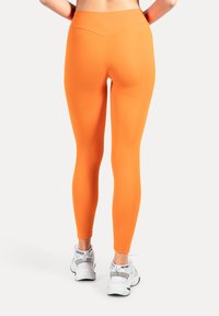 Leggings orange avec une taille haute, une coupe ajustée et une texture lisse, associées à des chaussures de sport blanches.