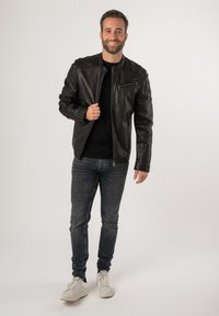 Giacca in pelle nera con design aderente, dotata di zip frontale e tasca sul petto, indossata sopra una camicia nera e abbinata a jeans scuri.