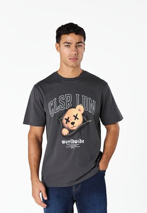 Jeune homme portant un t-shirt gris foncé avec un dessin d'ours aux yeux en forme de X et le texte "CLSR LDN" et "worldwide" sur le devant.