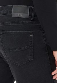 Jeans in denim neri con una patch in pelle nera texturizzata etichettata "Brax", con cuciture dettagliate e un classico design a cinque tasche.