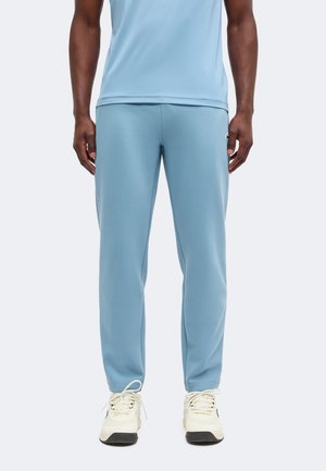 Homme portant un pantalon de survêtement bleu clair et une chemise assortie, associée à des baskets blanches, debout devant un fond uni.