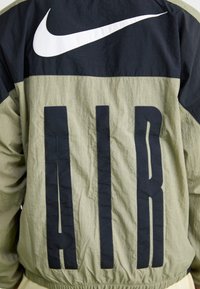 Nike vetrovka v olivno zeleni in mornarsko modri barvi, ki ima velik bel swoosh logotip in napis "AIR" v črni barvi na hrbtni strani. Teksturirana, lahka tkanina.