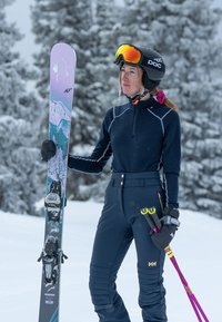 Narta z fioletowymi i turkusowymi grafikami, z czarnymi wiązaniami; narciarz z odpowiednim wyposażeniem, w granatowej koszulce z długim rękawem, dopasowanych granatowych spodniach i czarnych rękawicach.