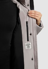 Hånd holder en åben lysegrå jakke, der viser sort indvendig foring, lodret lynlås-lomme og et stofmærke med teksten "Et varmt kram lavet af stof."