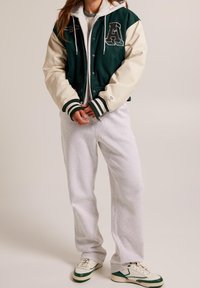 Vihreä ja kerma varsity-takki valkoisilla yksityiskohdilla, harmaa huppu, kengurutasku ja resorit hihansuissa. Yhdistetty harmaisiin collegehousuihin ja lenkkareihin.