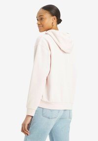 Sudadera de color rosa claro con cordón, bolsillo tipo canguro y puños acanalados. Llevada con jeans de color azul claro. La vista trasera muestra la capucha y el ajuste.