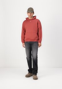 Red hoodie made of cotton, with a front pocket and minimal logo detail. Paired with dark blue jeans and beige shoes.