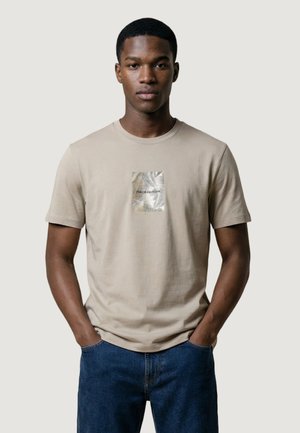 Młody mężczyzna w beżowej koszulce z małym kwadratowym, metalicznym wzorem liścia oraz ciemnoniebieskich dżinsach, stojący z rękami w kieszeniach.